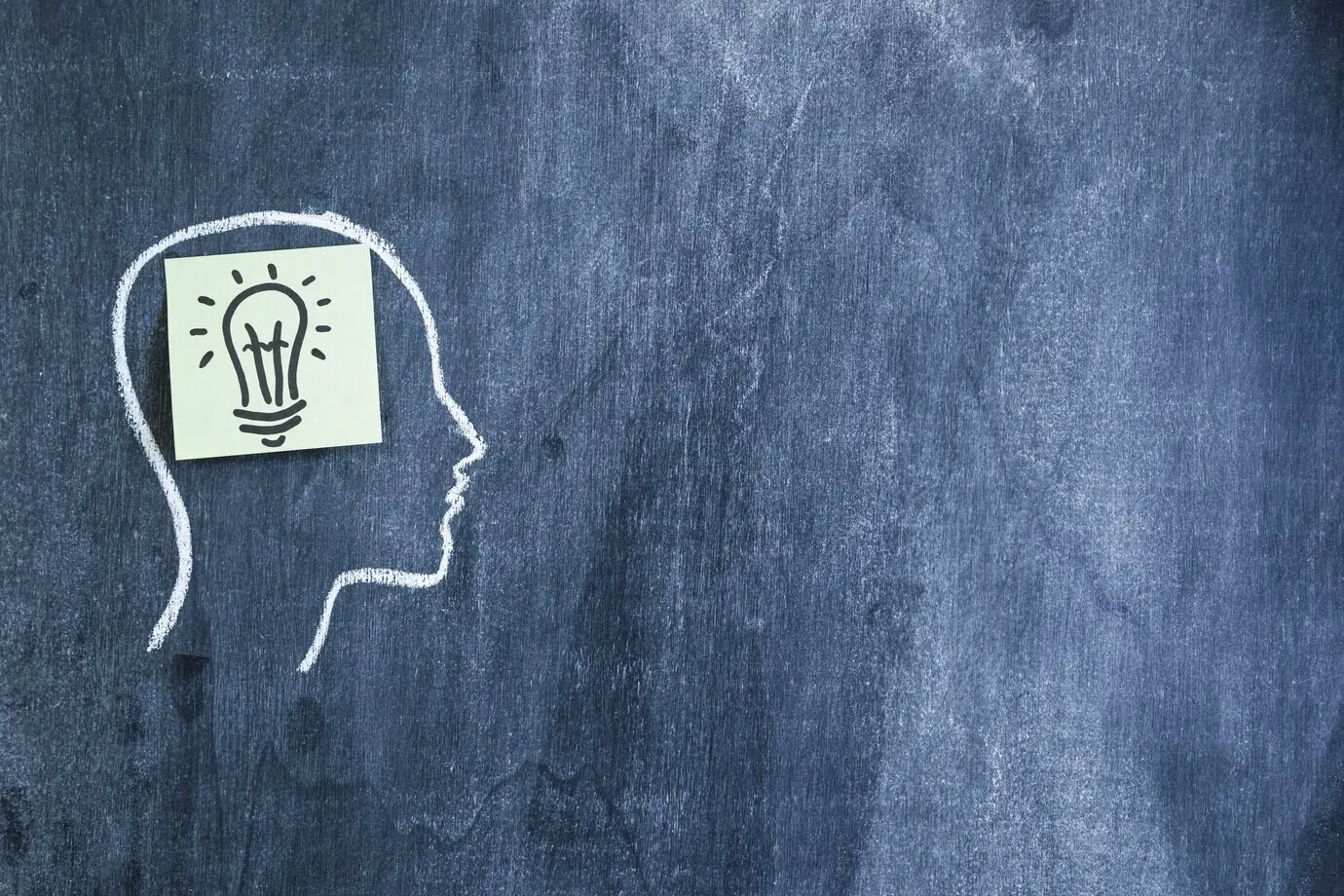 Липкая заметка «лампочка» на контуре лица, нарисованном мелом на черной доске.